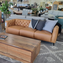 Tan leather 3 seater sofa with button-tufted backrest, styled in a showroom with neutral décor and modern furnishings.